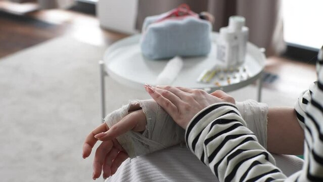 Close-up high-angle view of unrecognizable young woman with broken arm wrapped in white plaster bandage, gently massaging injured forearm sitting on couch by table with medicine at home, slow motion.
