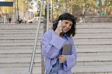 Cute charming lady with wonderful mile and short dark hairstyle wearing blue shirt is talking on smartphone, smiling and holding laptop in city square 