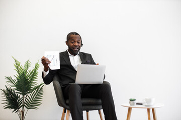 Competent african american financier presenting charts and graphs during online video meeting on modern laptop. Formally dressed man in headset sitting at office and having meeting on distance.