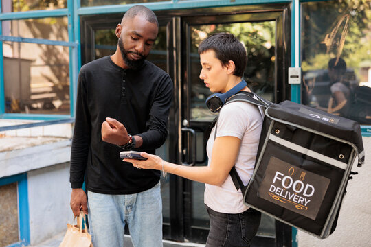 Client making contactless payment with smart watch for food delivery. Woman courier holding pos terminal device, customer paying for takeaway meal in front of office building medium shot