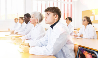 Fototapeta premium Young man in white coat sitting at desk in classroom, attending lecture in medical university with group of students and colleagues.