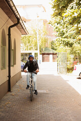 African american courier in headphones riding bike on street, looking at camera, front view. Deliveryman with customer order in backpack portrait, man delivering eatery meal