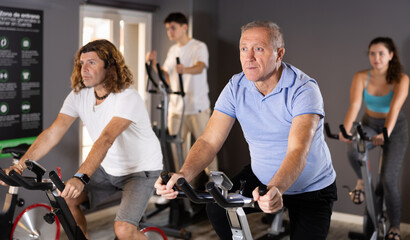 Fototapeta na wymiar Positive mature male riding exercise bike during cycling class in modern gym