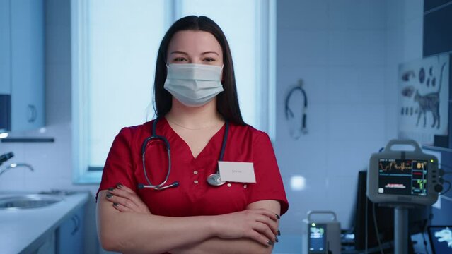 Close up of a female young veterinarian in a medical mask, who is crossing hands on chest and turning left and right, smiling at the camera. High quality 4k footage