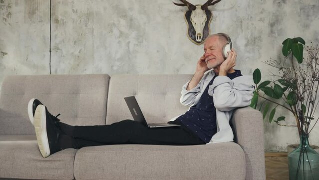 Elderly man listens to music on headphones, relaxes on the sofa at home. Enjoy music.