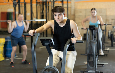 Dedicated active strong young man doing cardio workout on stationary bicycle as hard cross-training...