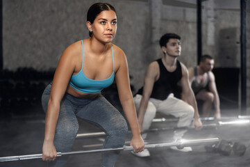 Motivated young woman practicing exercises with rod near other people in gym
