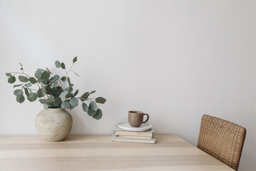 Silver green eucalyptus tree branches in beige textured vase. Brown cup of coffee, tea and old books on wooden table. White wall background. Minimalistic Scandinavian interior, dinning room. Breakfast - obrazy, fototapety, plakaty