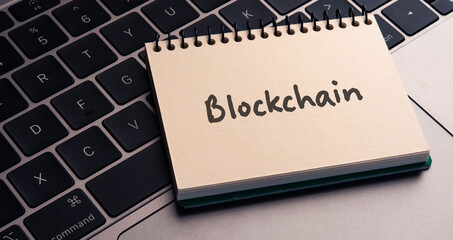 There is note book with the word Blockchain on a laptop. It is an eye-catching image.
