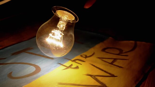 Male Reading a Text "Stop the war" on Ukrainian flag by Light of a Lamp in Dark. Drawing with lettering "Stop the war". Tungsten Incandescent lamp light bulb. Power outage. Blackout in Ukraine. War.