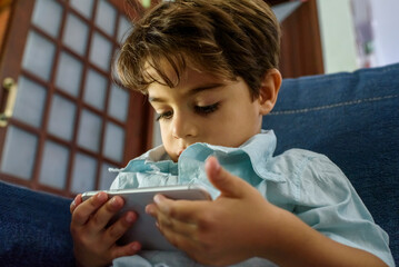 Boy about four years old watching video on mobile phone attentively on sofa at home.