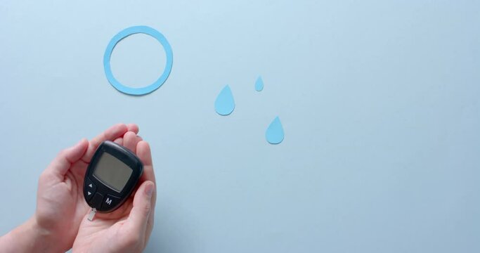Hands of caucasian woman holding glucometer over blue drops and circle on pale blue, slow motion