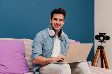 Handsome entrepreneur freelancer man working at home with a laptop sitting on a couch smiling and looking at camera. Young guy browsing on internet using a computer with wireless internet connection