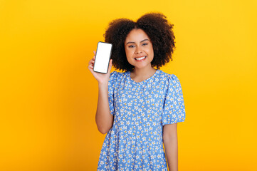 Joyful lovely african american or brazilian curly woman in summer dress, shows smart phone with mock-up empty white screen for advertising or presentation, smiles at camera, isolated orange background