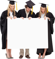 Three smiling students in graduate robe holding a blank sign