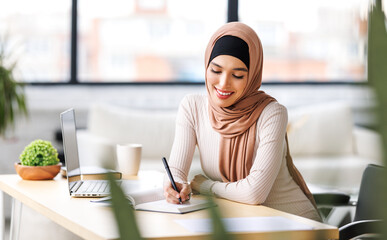 beautiful smiling muslim woman in traditional religious hijab works remotely on laptop from home