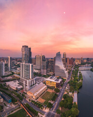 The photo captures the stunning Austin skyline during a beautiful sunset, with warm and vibrant colors illuminating the sky. The hazy atmosphere allows the moon to be visible, adding to the breathtak
