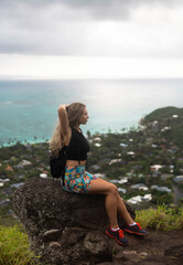 A woman hiker sits on the rock on top of a mountain. Hawaiian nature overcast cloudy weather.