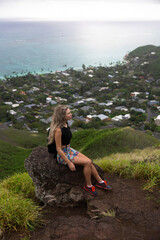 A woman hiker sits on the rock on top of a mountain. Hawaiian nature overcast cloudy weather.