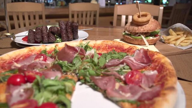 Static shot of a large meat pizza, burger, chips, and cevapcici served on a table
