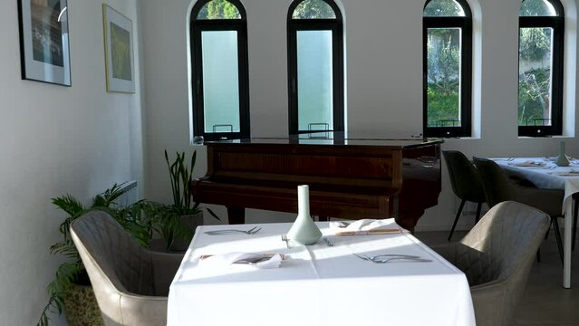 Fine dining room set with white tablecloths, silverware, vases, potted plants and a baby grand piano in the background.