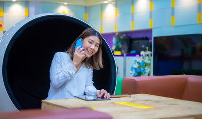 Beautiful asian business woman sitting on modern round chair and using phone to communicate with customer with smile, Digital marketing.