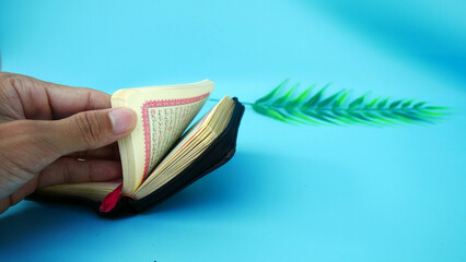 Muara Enim, Indonesia, April 04, 2023: Muslim man's left hand opens the Koran or Al Quran with copyspace cyan background. Quran hour and Nuzul Quran concept.