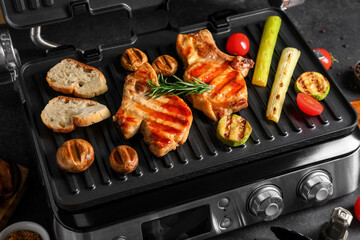 Modern electric grill with tasty steaks and vegetables on dark background