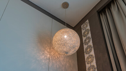 A decorative lamp is suspended on the ceiling, in the corner of the room. Braided lampshade in the form of a ball. The lights are on.