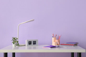 Workplace with stationery, clock and lamp near lilac wall