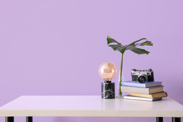 Photo camera with books, palm leaf and lamp on table near lilac wall