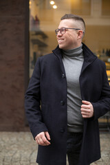 Positive emotions. Attractive businessman in glasses, dressed in gray sweater and black cashmere coat on city street.