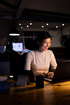 Happy asian businesswoman sitting at desk, using laptop and working late at office