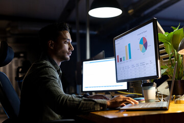 Biracial businessman sitting at desk, using computers and laptop, working late at office