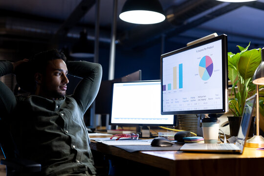 Biracial businessman sitting at desk, using computers and laptop, working late at office