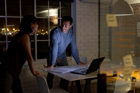 Diverse business people standing at desk, using laptop and discussing, working late at office