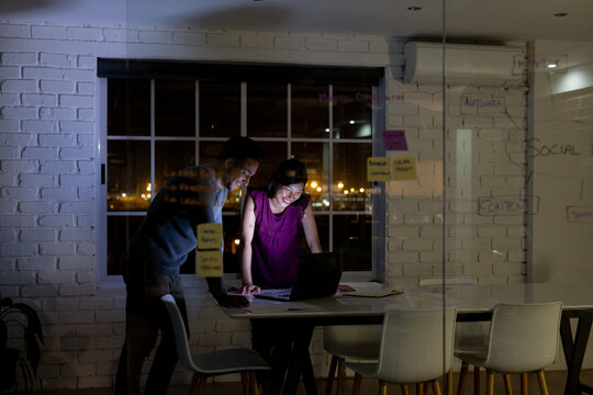 Diverse business people standing at desk, using laptop and discussing, working late at office