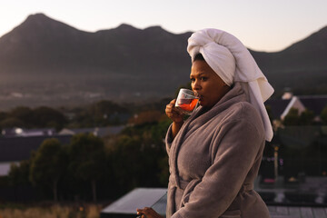 Happy plus size african american woman wearing robe and towel on head, drinking tea on balcony