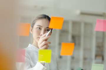 Asian woman working on project plan using sticky papers notes on glass wall, people meeting to share idea, Business design planning concepts.