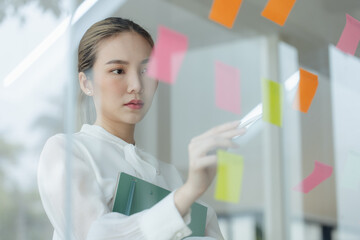 Asian woman working on project plan using sticky papers notes on glass wall, people meeting to share idea, Business design planning concepts.