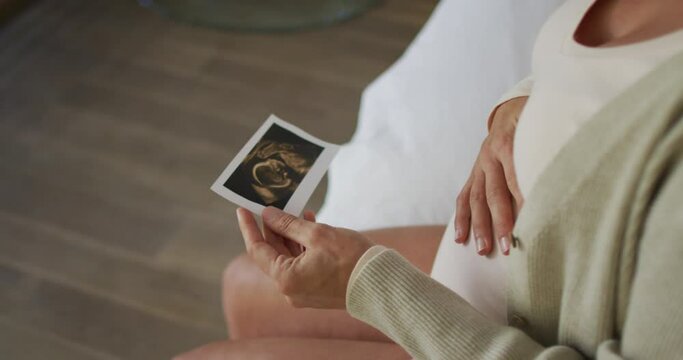 Hands of caucasian pregnant woman sitting on bed, touching belly and looking on ustrasound photo