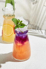 Violet and mango lemonade in jug on white background. Colorful lemonade with violet syrup and citrus fresh in minimal style. Summer drink with palm shadows. Mocktail on concrete wall.