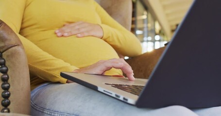 Midsection of caucasian pregnant woman sitting in armchair with laptop