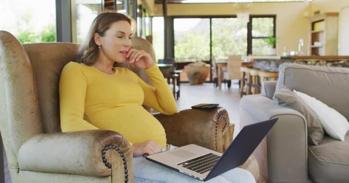 Happy caucasian pregnant woman sitting in armchair and using laptop