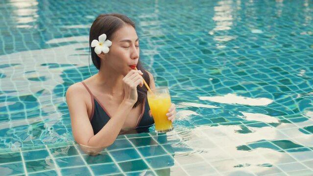Youth attractive Asia beauty female wear bikini stand in swimming pool use straw drink cocktail enjoy weekend nice view relax nature at hotel villa on vacation. Real estate property facility concept.