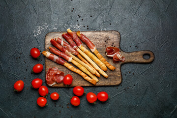 Wooden board of tasty Italian Grissini with bacon and tomatoes on dark background