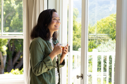 Happy asian woman looking through window, smiling and drinking tea