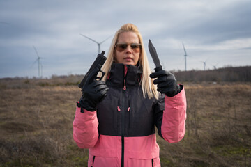 A girl in sunglasses with a gun and a knife in her hands in nature near wind generators.