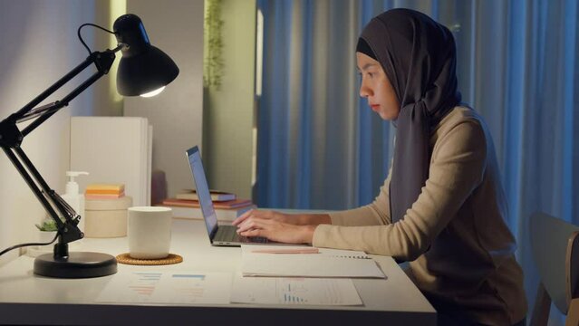 Young happy Asia muslim freelancer woman wear hijab casual clothing typing keyboard laptop computer at home office overtime working at night. Small business owner online marketing e-commerce concept.