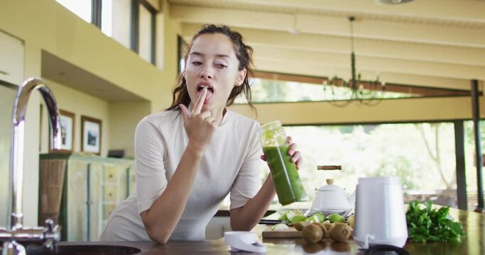 Happy biracial woman with vitiligo tasting smoothie from fingers in kitchen